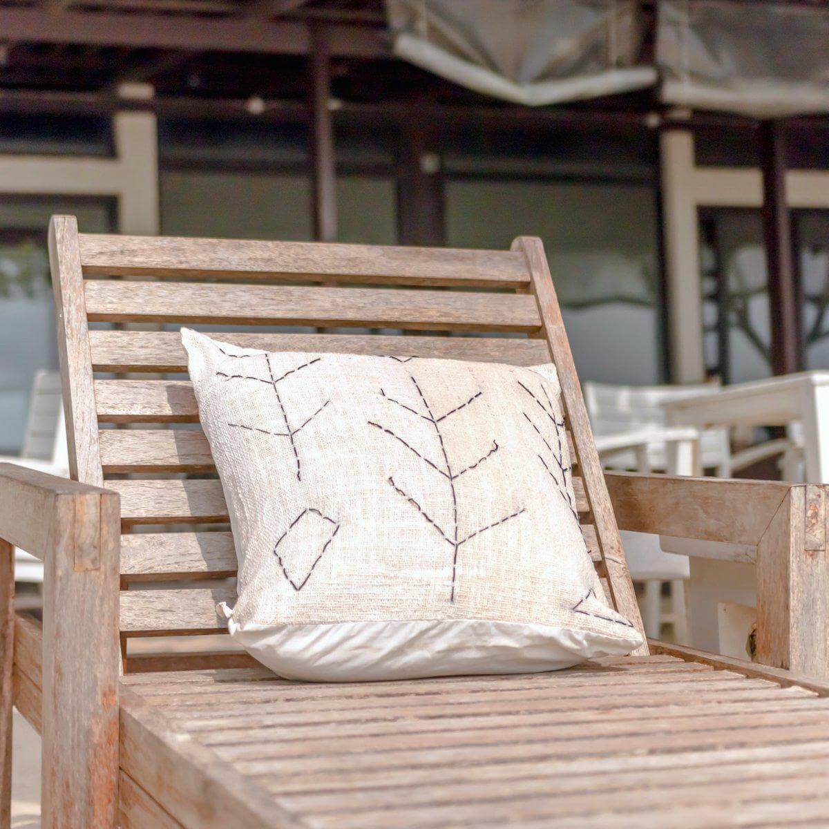 Alt Tag: Handgeweven Boho-kussen SERAJA op houten stoel in zonnige tuin, natuurlijke katoen stof, perfect voor decoratieve touch.