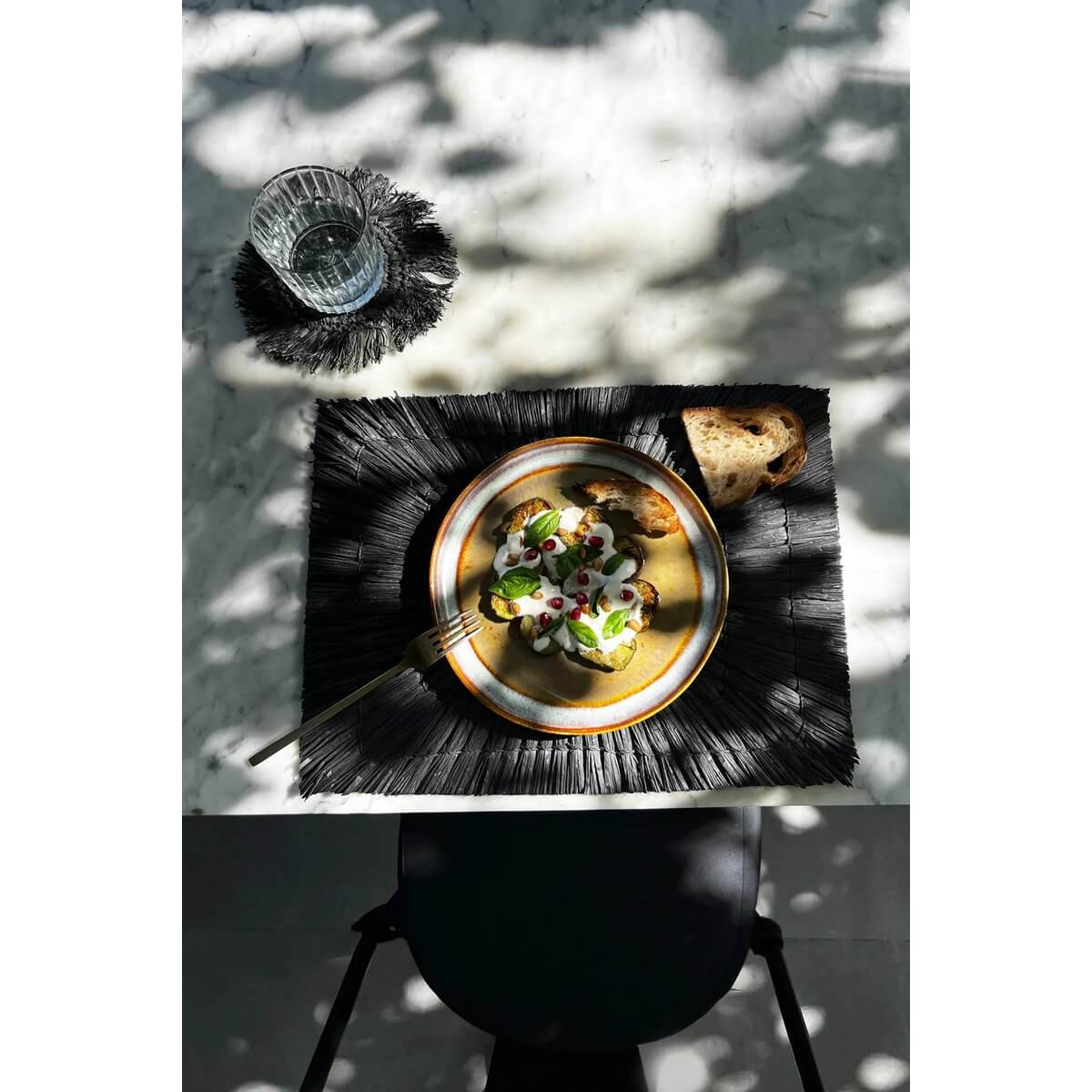 Bohemian table setting with black De Alang Alang Placemat featuring a dish and glass under dappled sunlight.