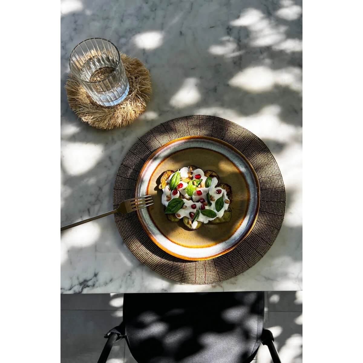 Bohemian table setting with a De Beaded Placemat in gold and a plate of salad, under dappled sunlight on a marble surface.