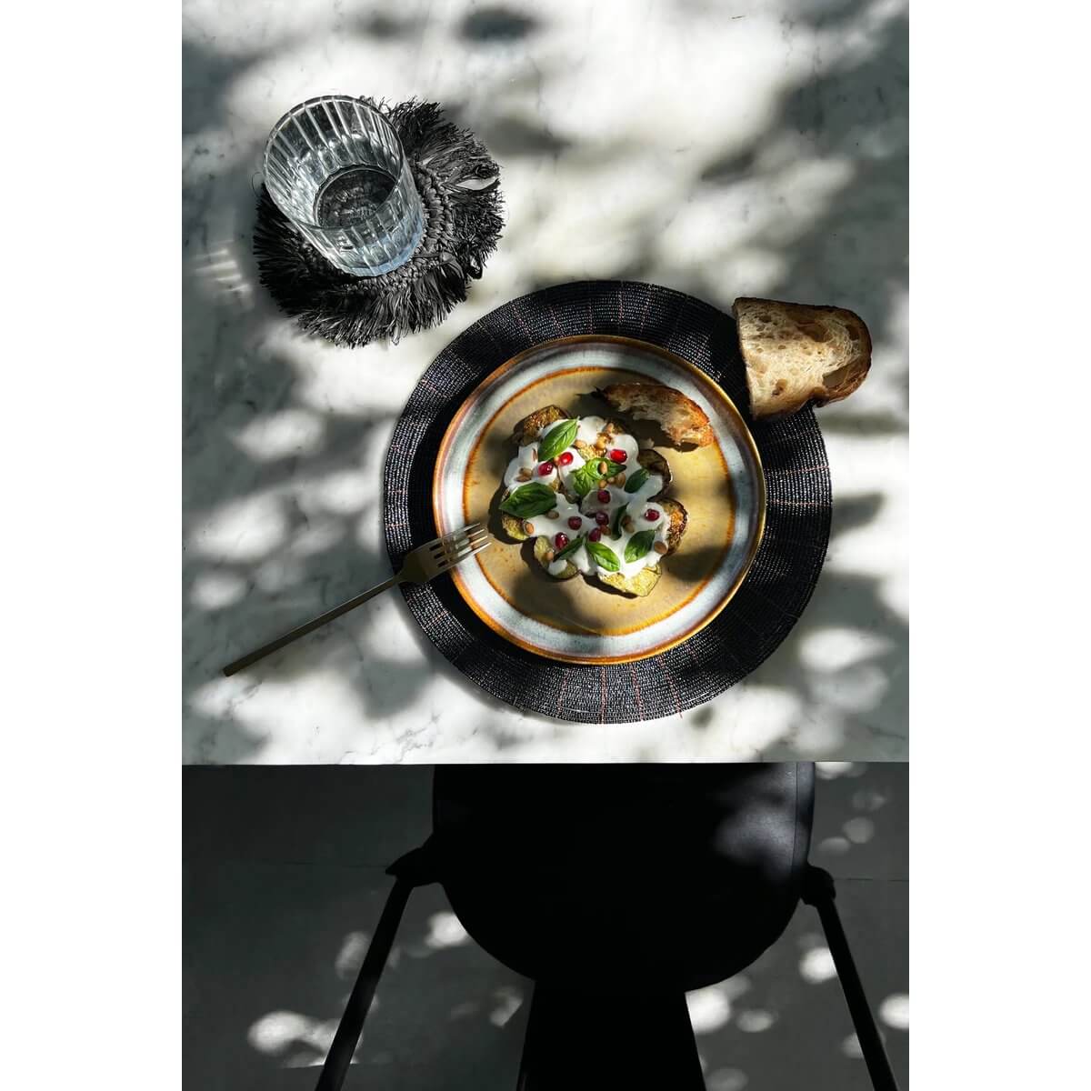 Bohemian chic table setting with De Beaded Placemat - Zwart, topped with a gourmet dish and rustic bread, under dappled sunlight.