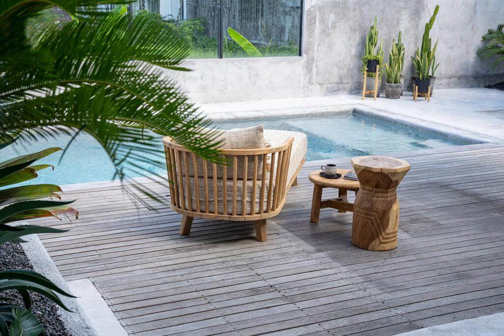 Houten De Bloom Kruk naast een zwembad op een terras, omgeven door groene palmbladeren en moderne decoratie, straalt rust en stijl uit.