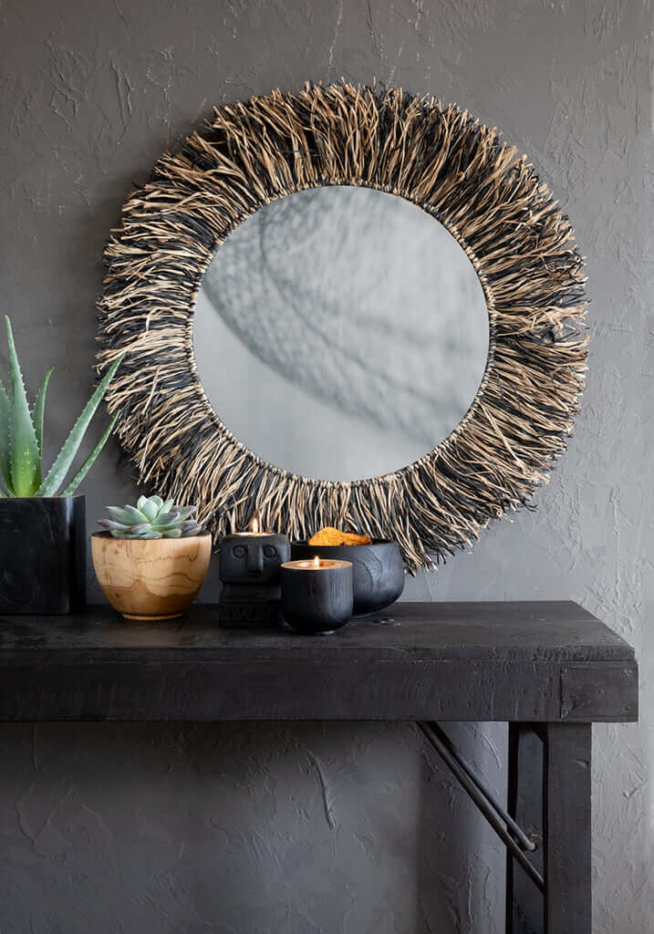 Wandspiegel met gevlochten raffia omlijsting boven een houten tafel met decoratieve planten en accessoires.
