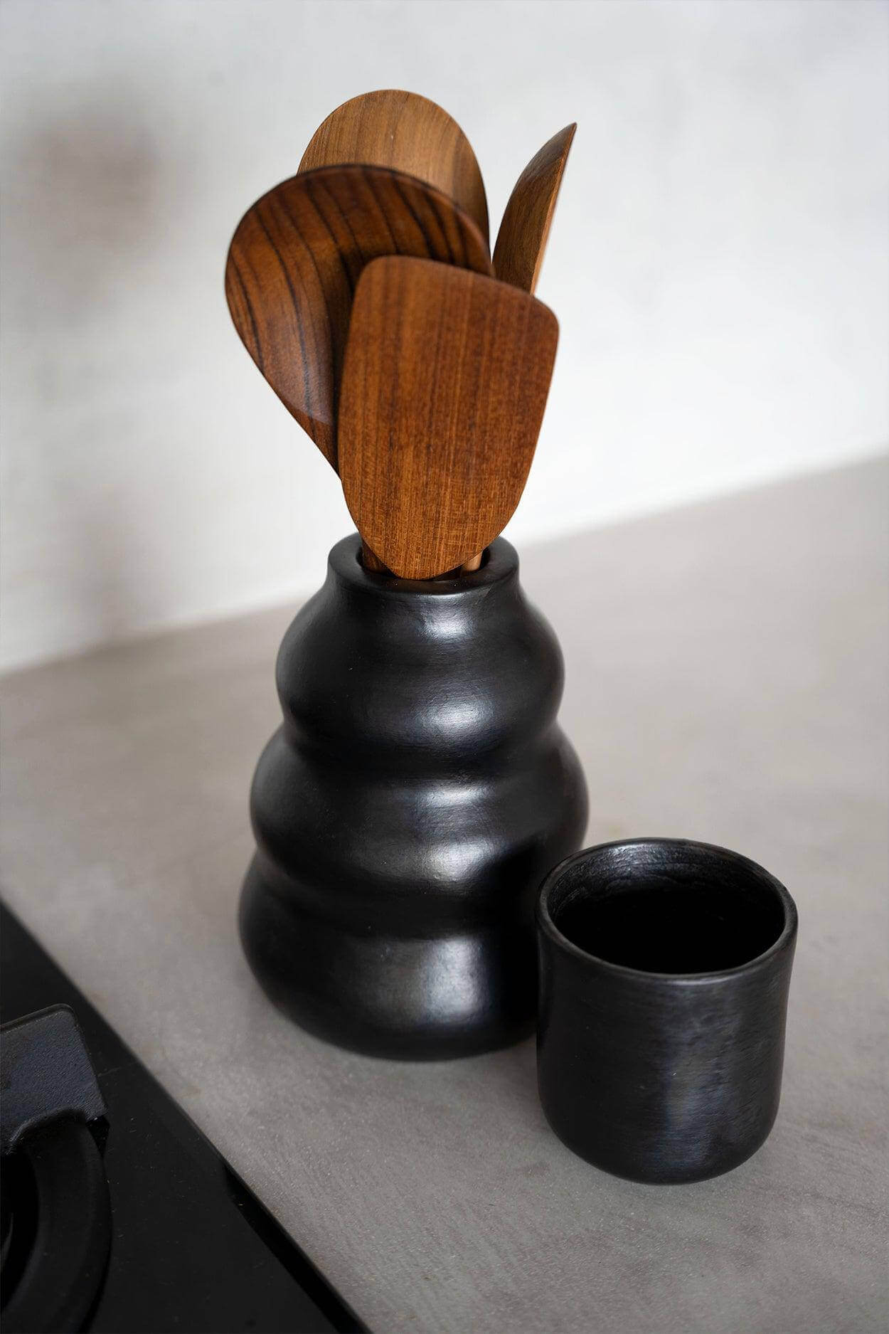 Zwarte keramische beker uit de Burned collectie met houten lepels op een grijs aanrecht, perfect voor kleurrijke maaltijden.