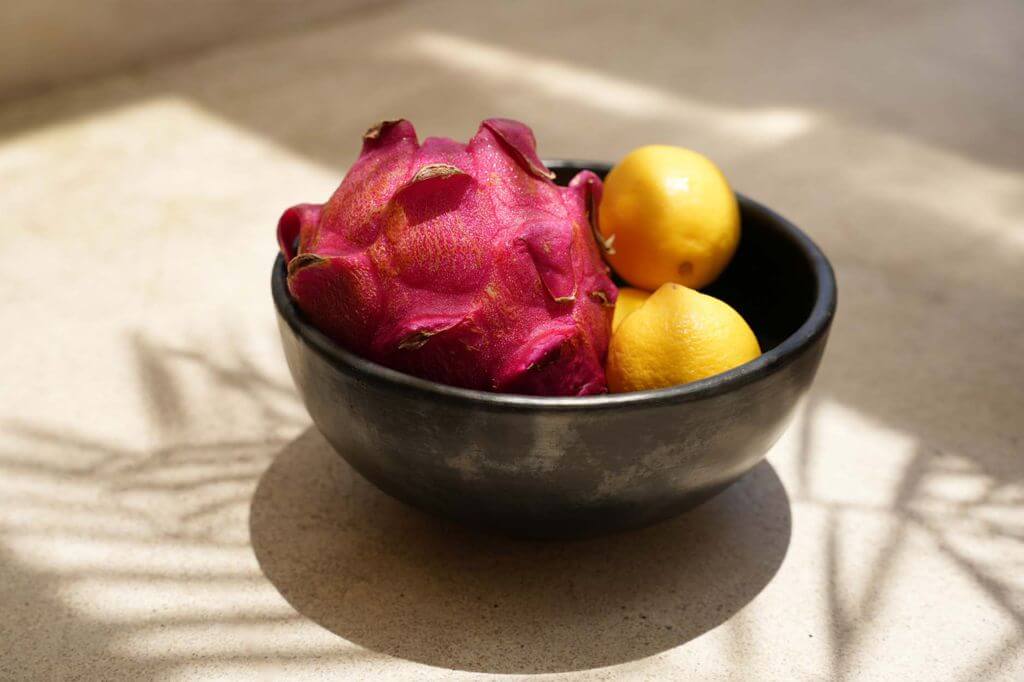 Zwarte gebrande terracotta kom met een pitaya en twee citroenen, op een zonovergoten tafel, belicht het unieke servies design.