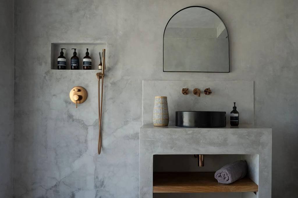 Stijlvolle badkamer met betonnen muren, gouden kranen en een terracotta vaas met rotan detail voor een kustsfeer.