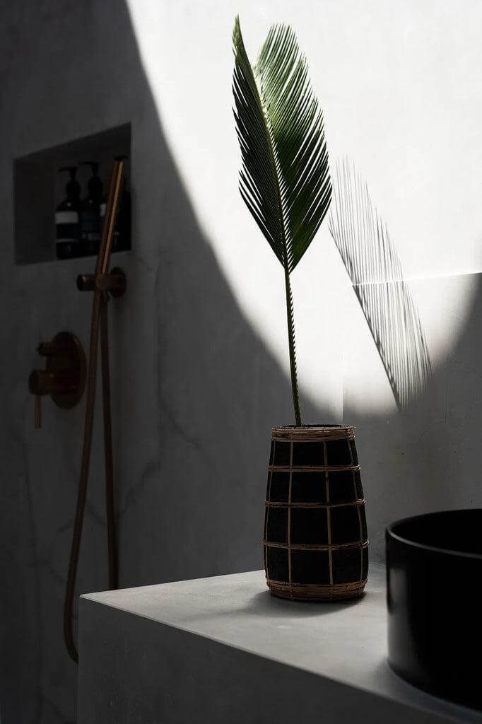 Stijlvolle zwarte terracotta vaas met rotan detail en droog palmblad in een elegante badkameromgeving.