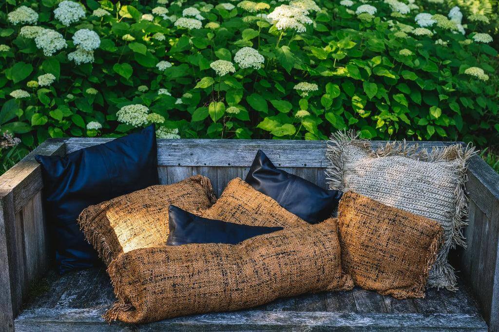 Zwarte lederen en bruine stoffen kussens op een houten bankje met groene planten op de achtergrond, buitenlounge setting.