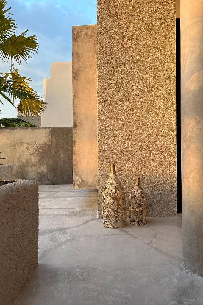 Rotan De Garaffa hanglampen in flesvorm op betonnen vloer, omgeven door moderne architectuur en palmboom, bij zonsondergang.