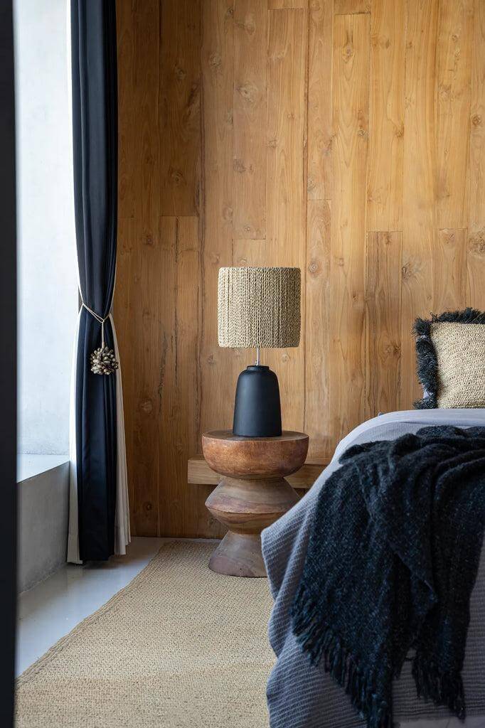 Handgemaakte suarhouten kruk in natuurkleur als bijzettafel, met designlamp, tegen een houten muur naast bed. Perfect voor moderne interieurs.