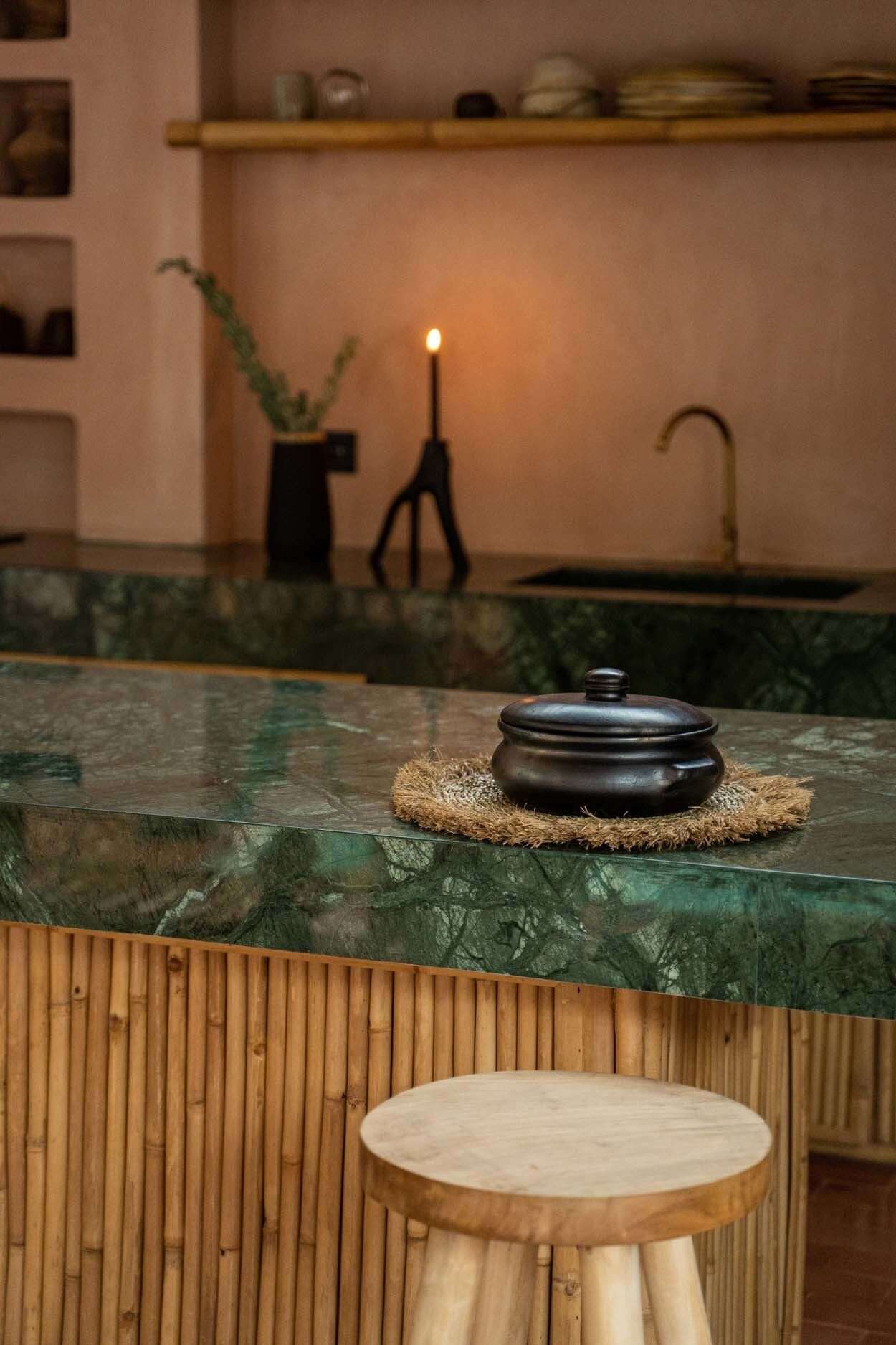Gezellige keuken met bamboe eiland, groene marmeren werkblad en zwarte pan, met een barstoel op de voorgrond voor een huiselijke sfeer.