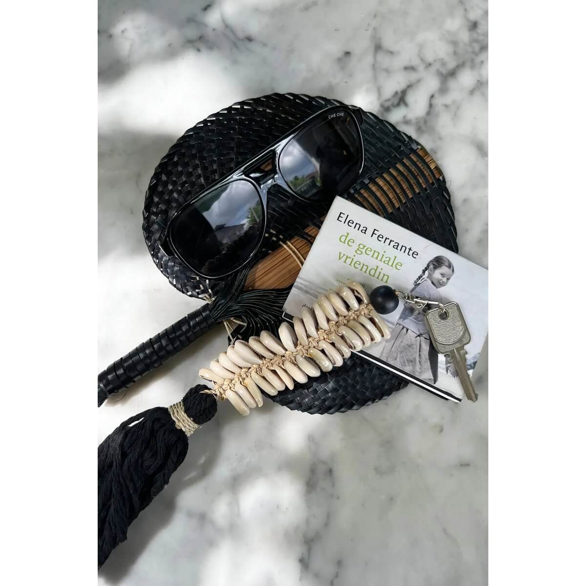 Bohemian macrame fishbone keychain with black tassel and shells, styled with sunglasses and a book on a marble surface.