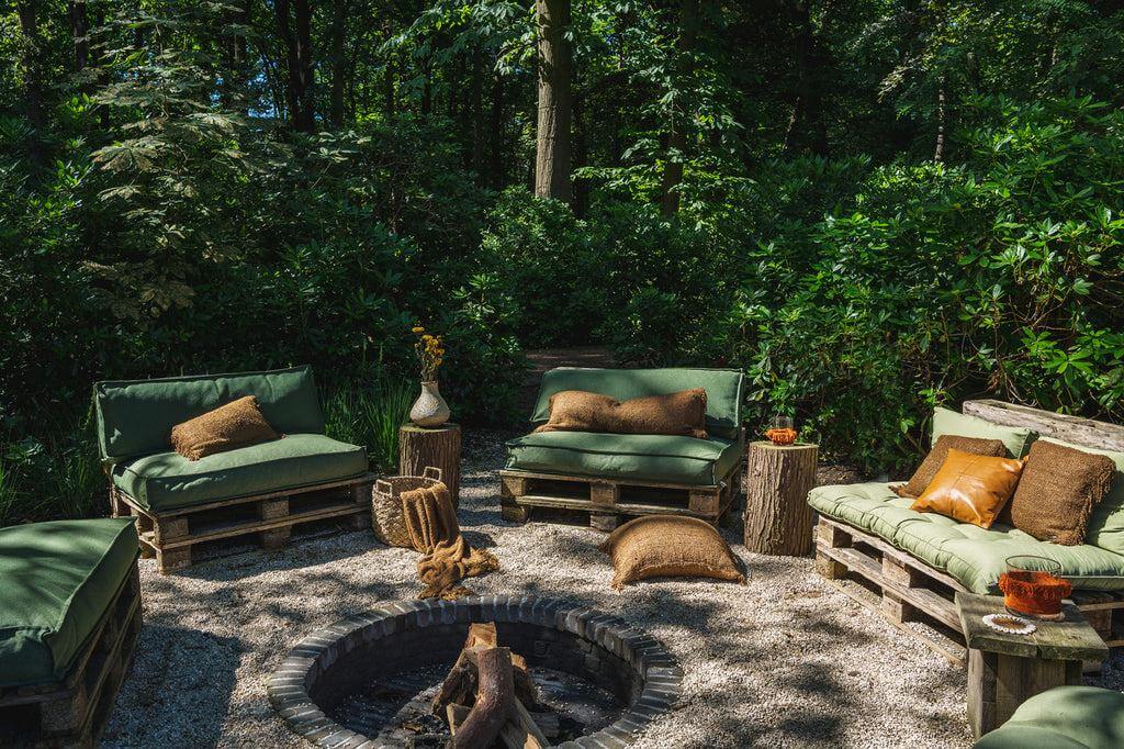 Boho tuinsetting met groene pallets en bruine kussens, omringd door weelderige bomen, met vuurplaats centraal en natuurlijke decor elementen.