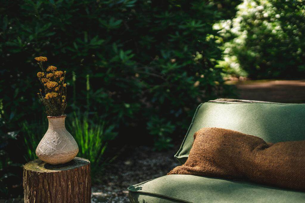 Gezellige buitenplek met groene bank en boho bruine kussenhoes, naast een vaas met gele bloemen op een houten krukje.