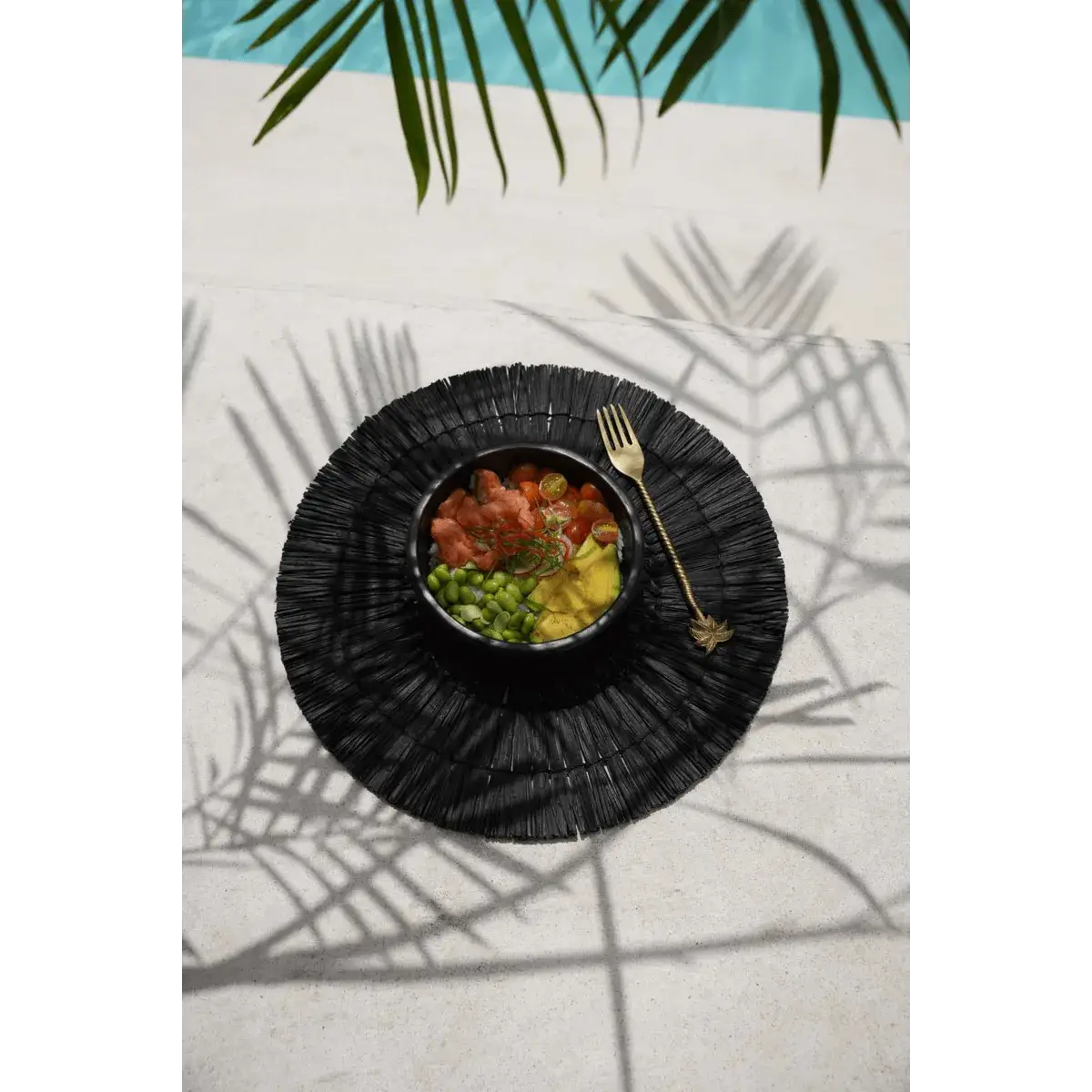 Bohemian-style black placemat with a luxurious gold palm tree fork beside a vibrant tropical salad, exuding elegance by the poolside.