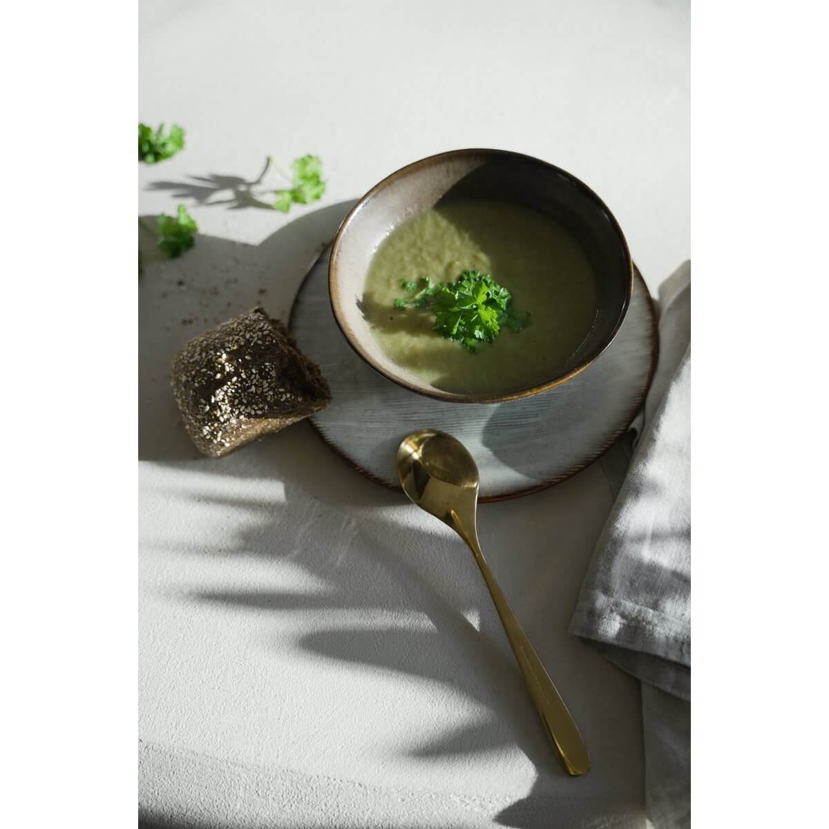 De Peniche cereal bowl with soup, garnished with parsley, accompanied by crusty bread and gold spoon, showcasing elegant table setting.