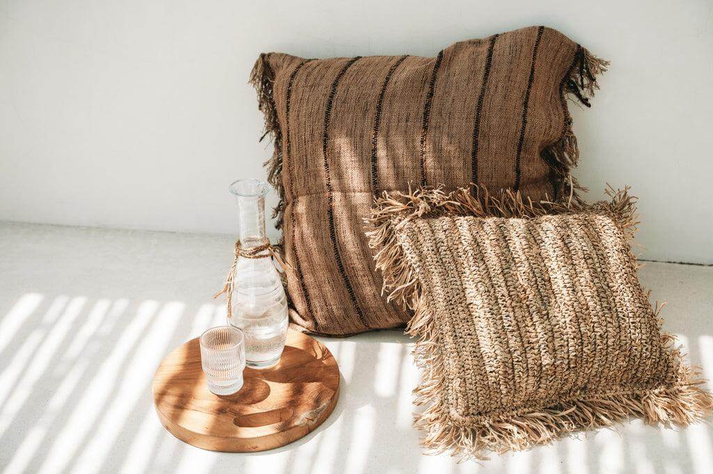 Alt Tag: Handgeweven raffia kussenhoes in natuurlijke tinten op een bank, met boho-vibe en zonnige uitstraling. Perfect voor huisdecor.