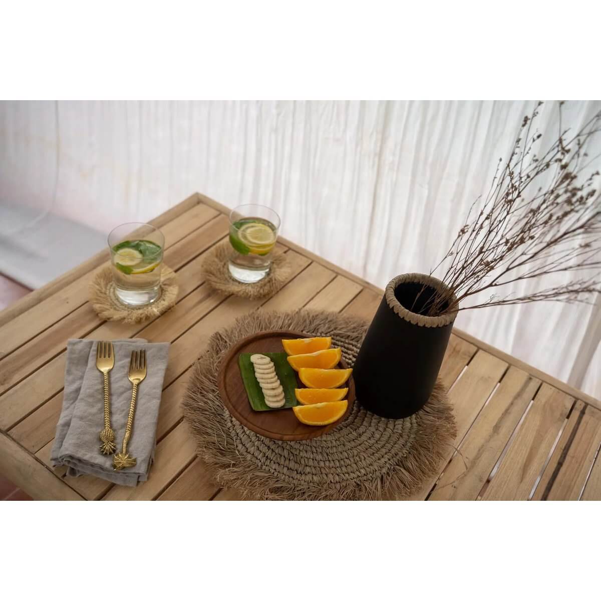 Cozy bohemian table setting with De Seagrass Raffia Placemat, citrus slices on a wooden tray, drinks, and cutlery for tropical flair.