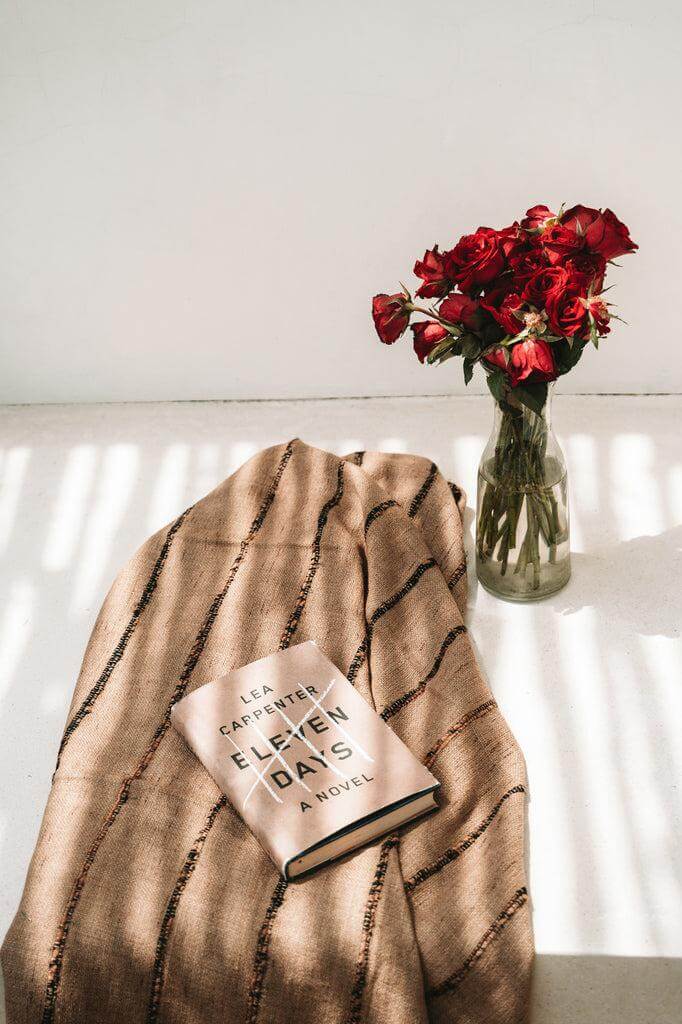 Alt Tag: Elegante handgeweven plaid in beige en zwart naast een boek en bloeiende rode bloemen in vaas, perfect voor gezellige avonden.