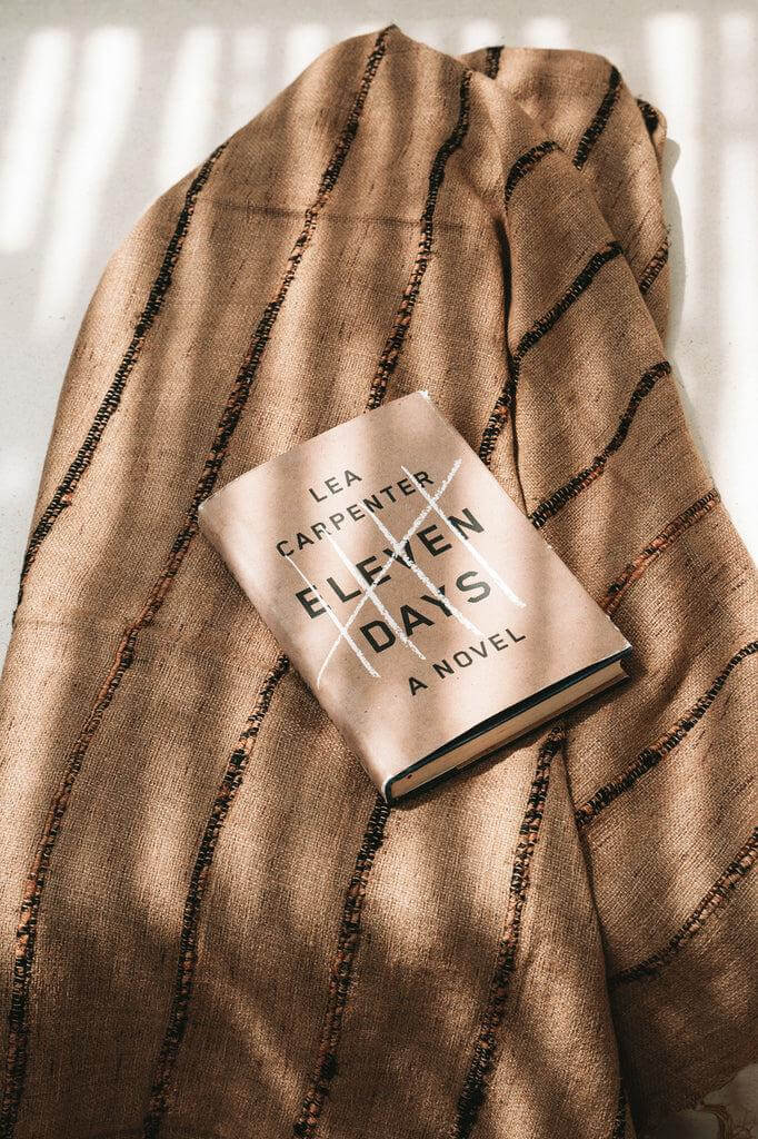 Luxe handgeweven katoenen plaid in beige en zwart, met een roman op de plaid geplaatst, tegen een warme setting. Perfect voor knusse avonden.