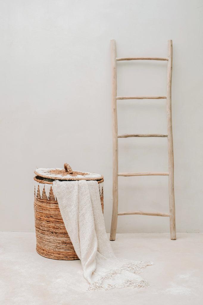 Luxe handgeweven katoenen plaid in crème met franjes, elegant gedrapeerd over een stijlvolle mand naast een houten ladder.