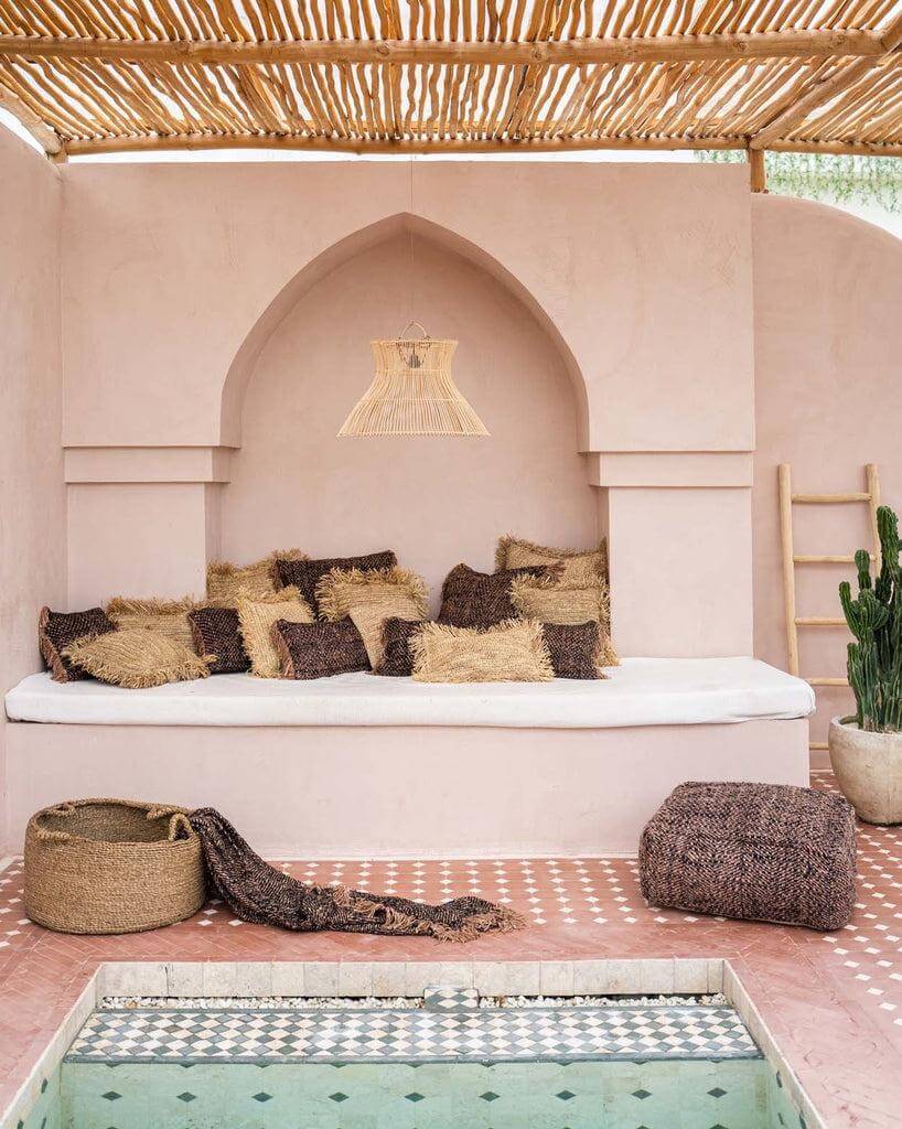 Bohemien patio met beige bank, kussens en decoratieve lamp, naast een zwembad en groene cactus onder een houten overkapping.