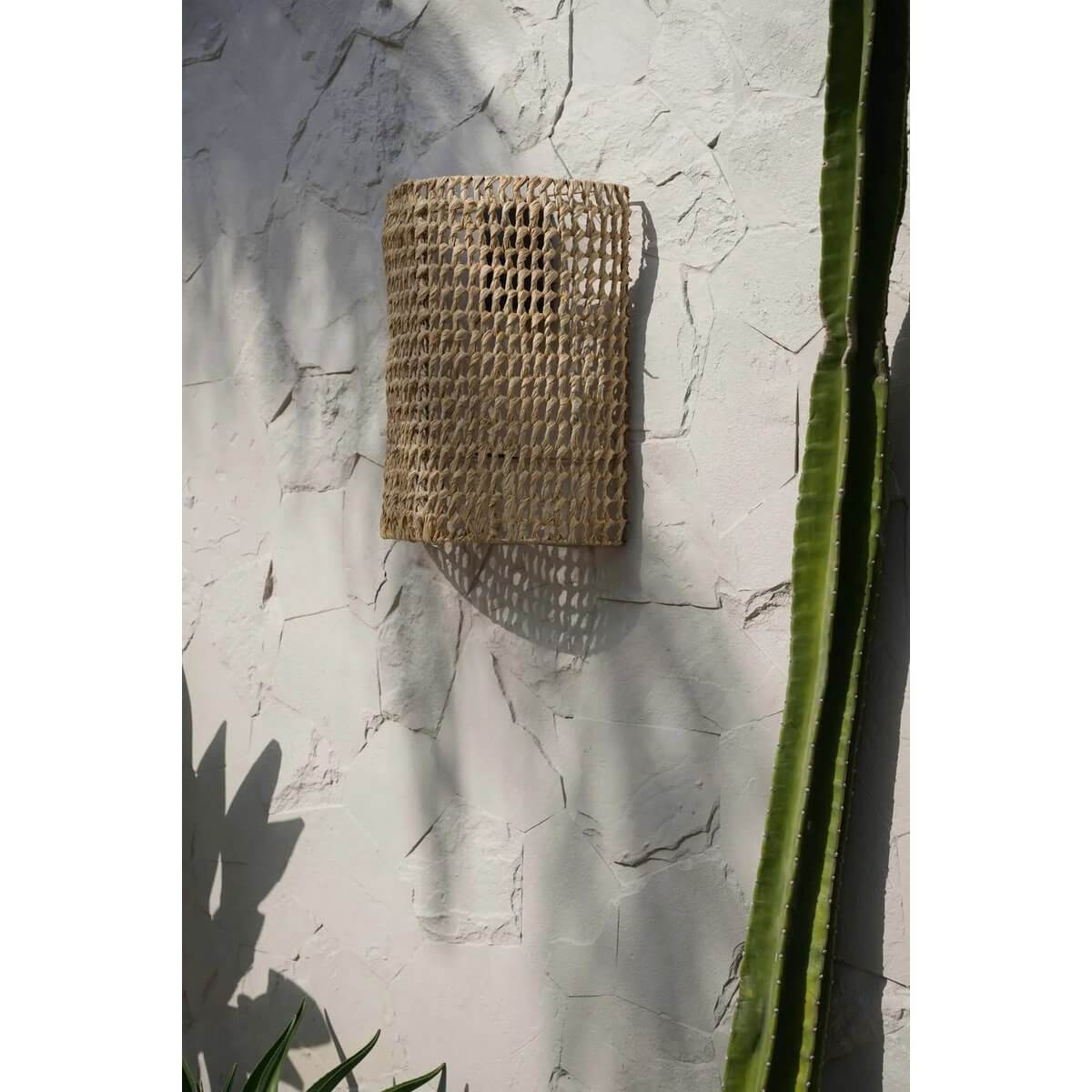 De SiriKit Wandappliqué in natuurlijk sisalweefsel, straalt warme gloed uit op witte muur met cactus.