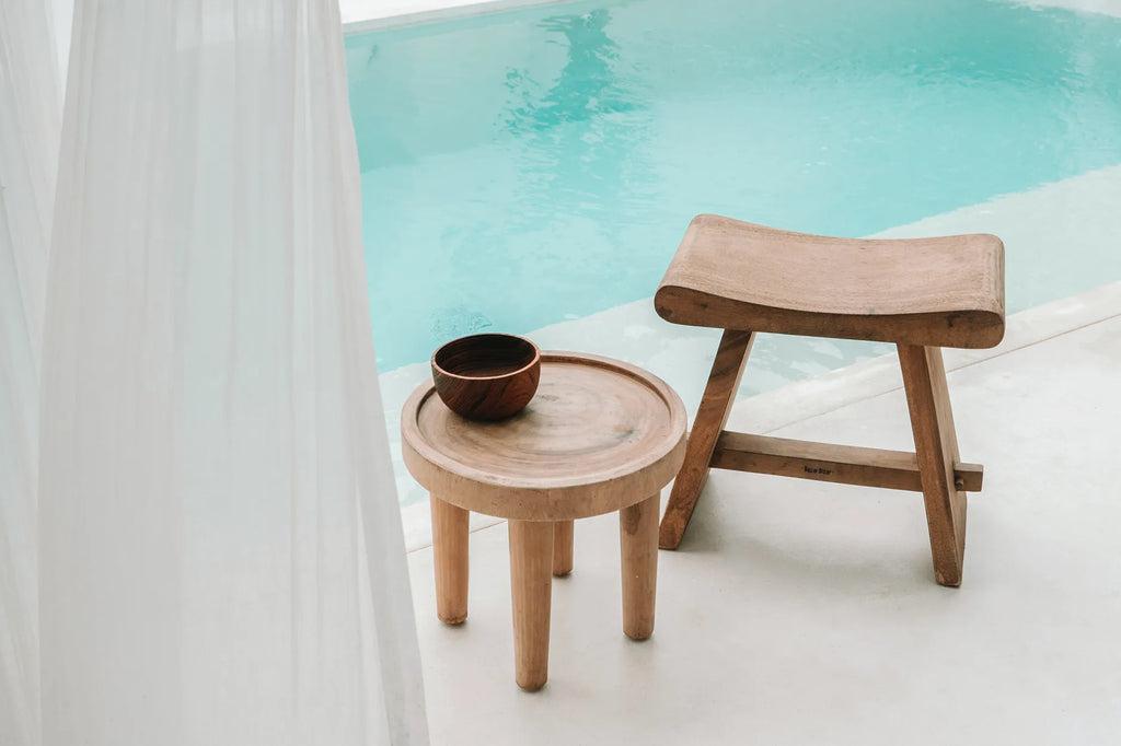 Houten De Suar kruk en tafel naast een zwembad, ideaal voor binnen en buiten decoratie, met een natuurlijke en klassieke uitstraling.