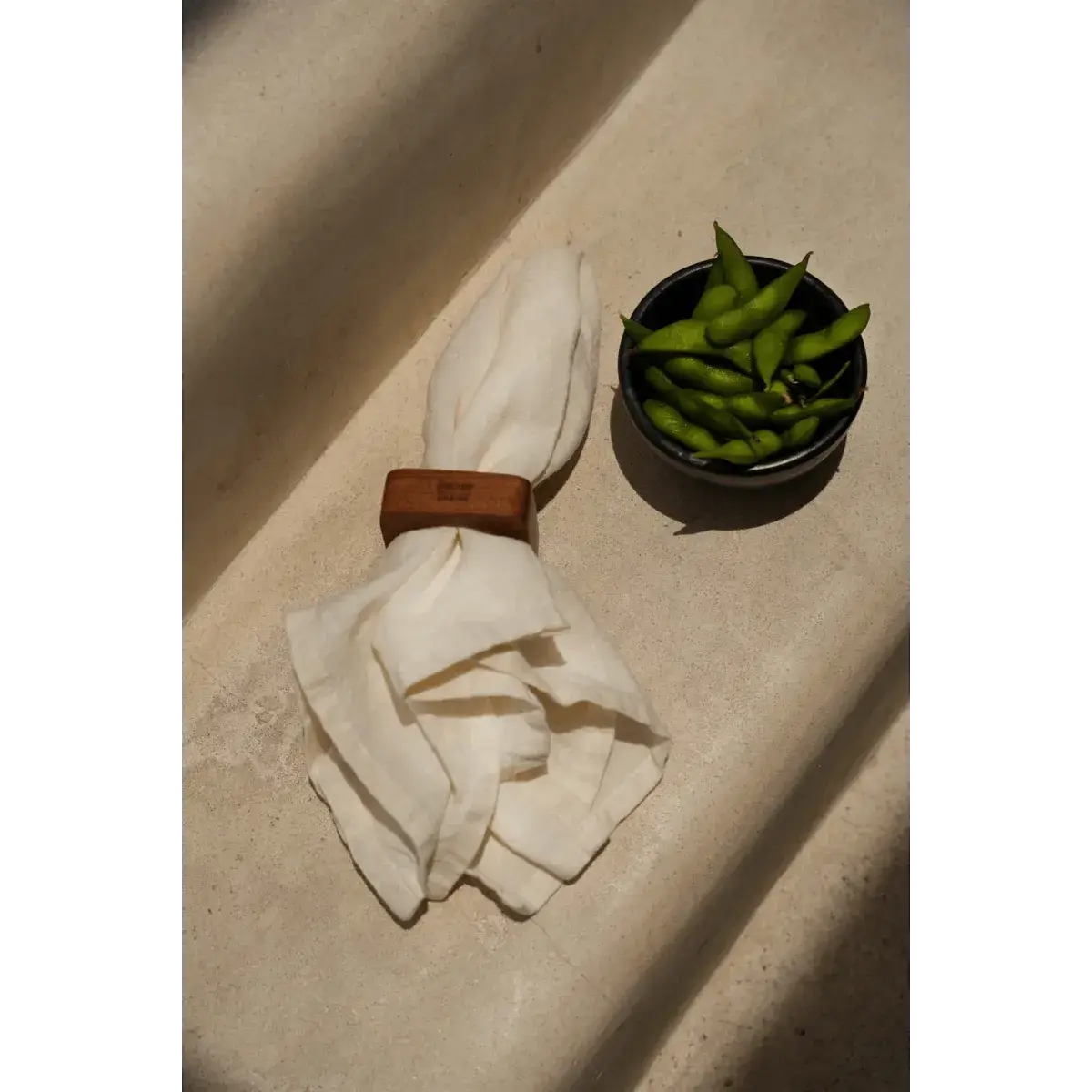 De Teak Root Servetring houdt een witte servet vast naast een schaaltje groene peulen, perfect voor bohemian eetgelegenheden.