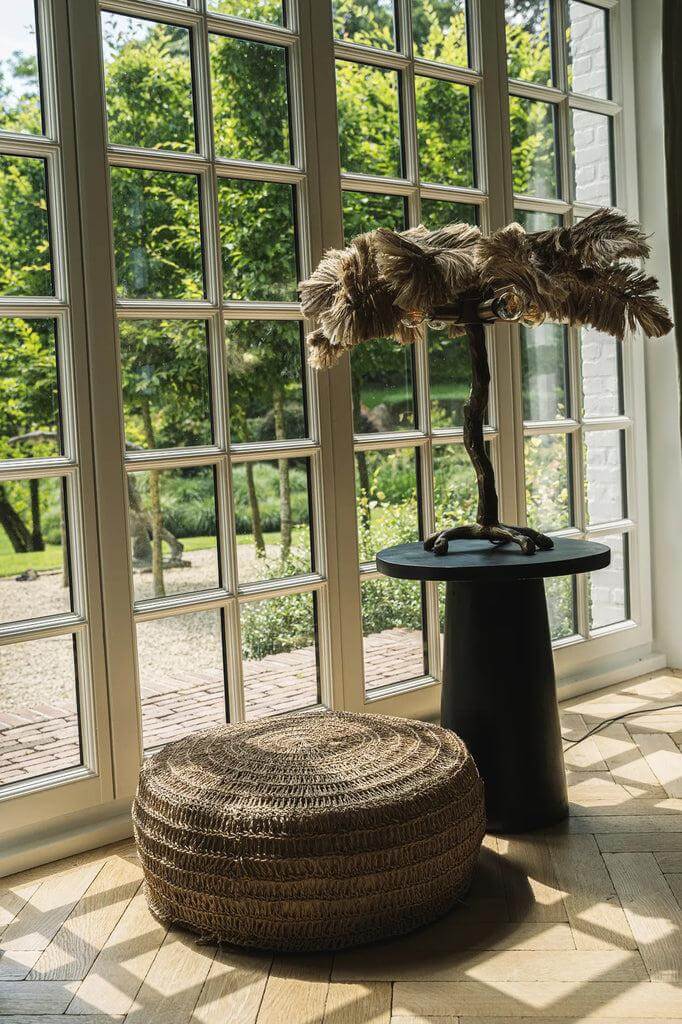 De Timber Conic Bijzettafel van zwart Munggur hout naast een rieten poef bij een raam. Natuurinspiratie in huis met unieke patronen.