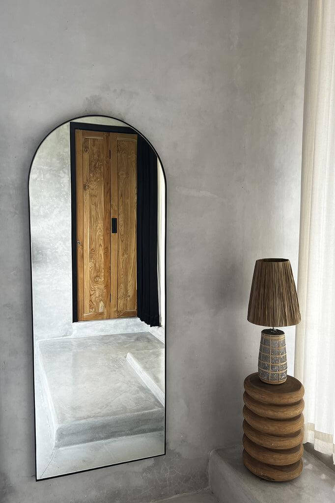 Stijlvolle hal met een intieme sfeer, voorzien van een vloerlamp met houten ringen naast een hoge, moderne spiegel met houten deurreflectie.