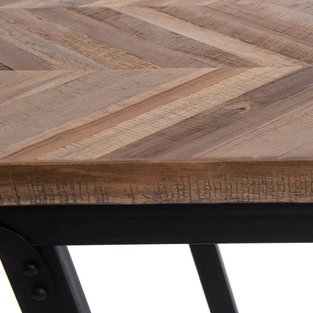 Detail van een teakhouten tafel met visgraatpatroon op zwarte ijzeren poten, geschikt als eettafel voor binnen of overdekt buiten.