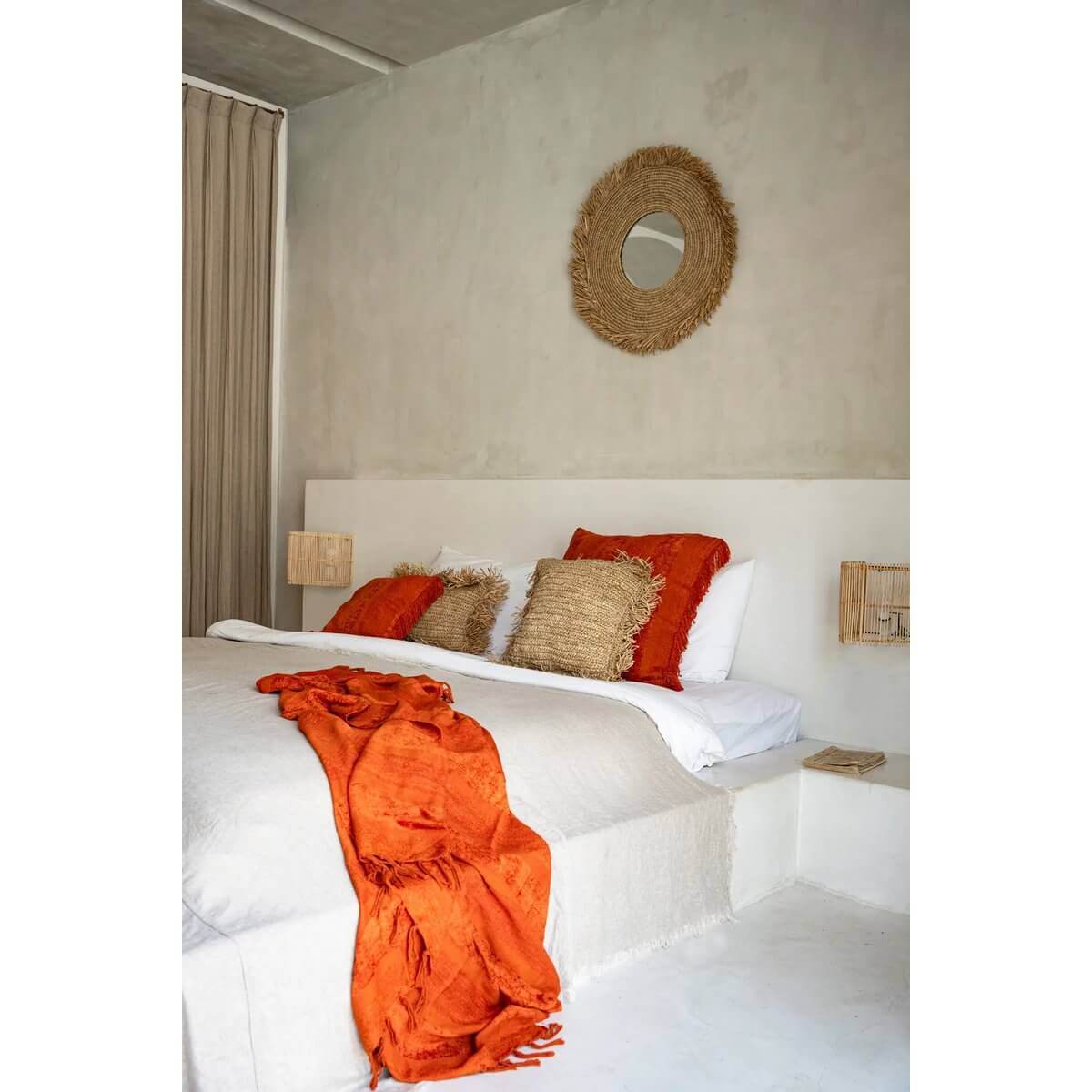 Sfeervolle slaapkamer met De Yema Spiegel aan de muur, geweven raffia accenten en warme oranje kussens voor een rustieke uitstraling.