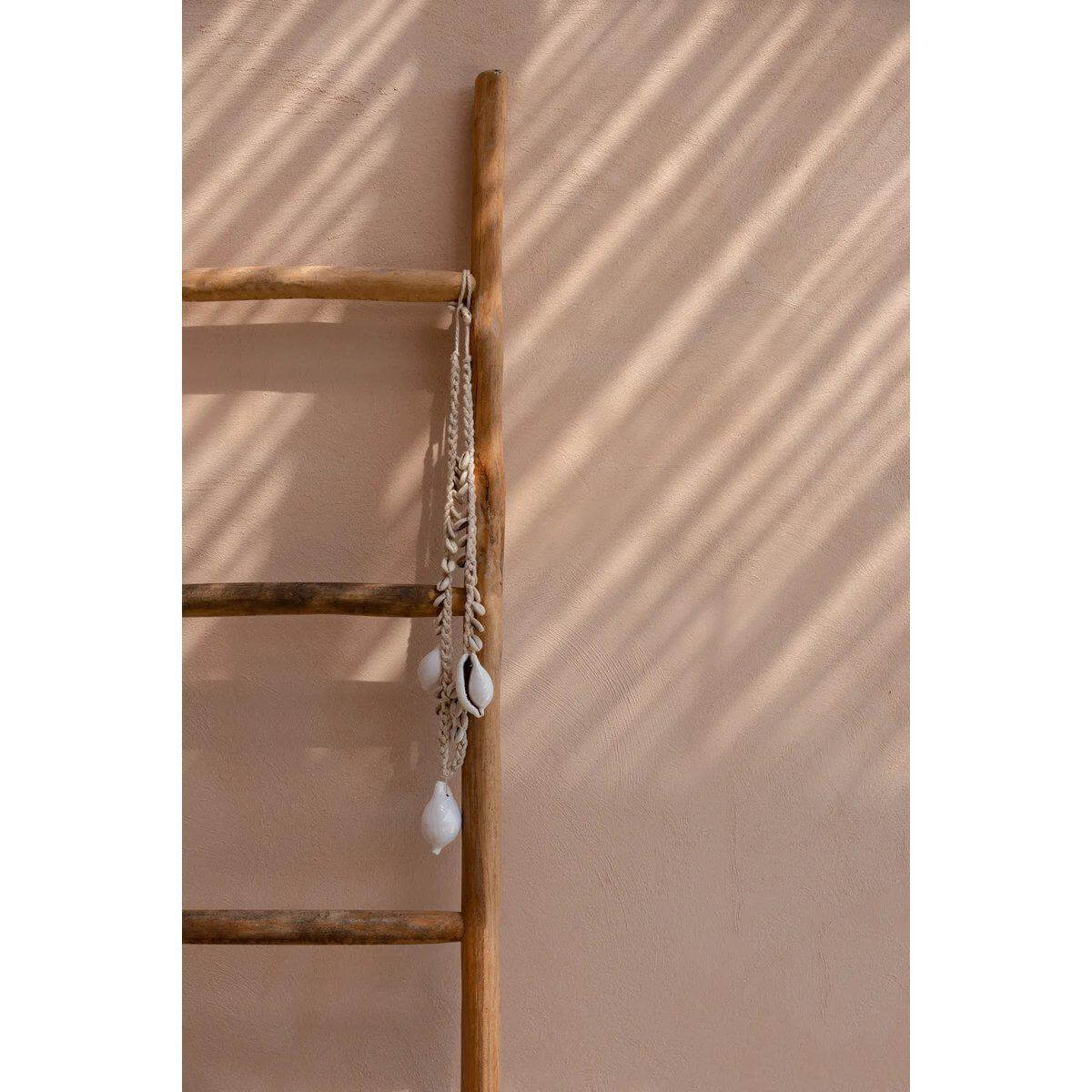 De grote witte kauri schelp ketting hangt sierlijk aan een houten ladder tegen een beige muur met zonnestralen. Kust en bohemien stijl.