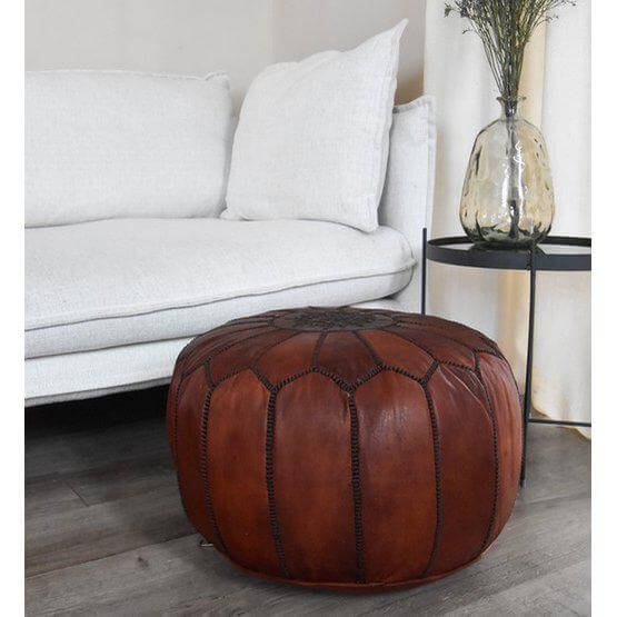 Stijlvolle kaneelbruine leren poef naast een moderne witte bank en glazen vaas, perfect voor boho decor en multifunctioneel gebruik.