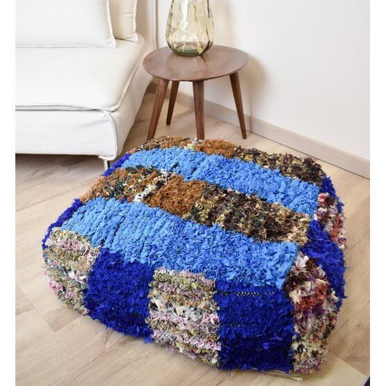 Marokkaans Boucharouite vloerkussen in blauwe en aardetinten, handgemaakt van gerecycled textiel, opgelicht naast stoeltje.