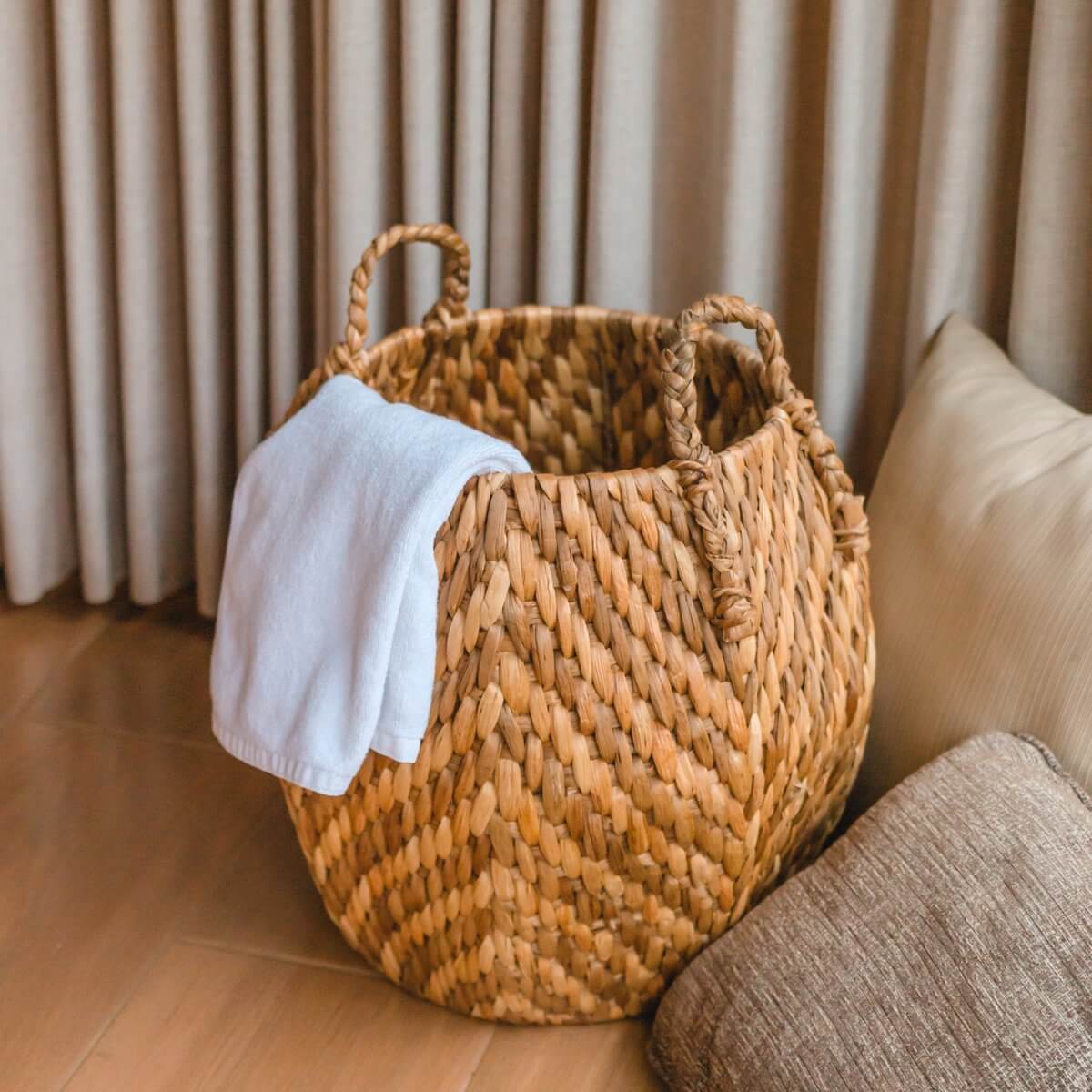 Opbergmand WAYAG van waterhyacint met handvatten, ideaal als wasmand of plantenhouder, naast beige kussens.