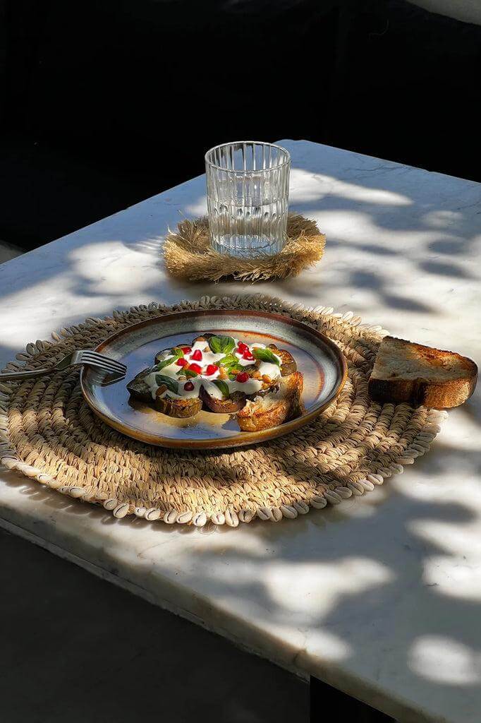 Elegante placemat van raffia met kauri schelpen, perfect voor boho stijl tafelsetting. Compleet met glas en smakelijke toast op bord.