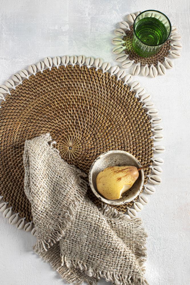 Handgeweven rotan placemat omgeven door kauri schelpen met een rustieke servet, een kom en een glas op een tafel.