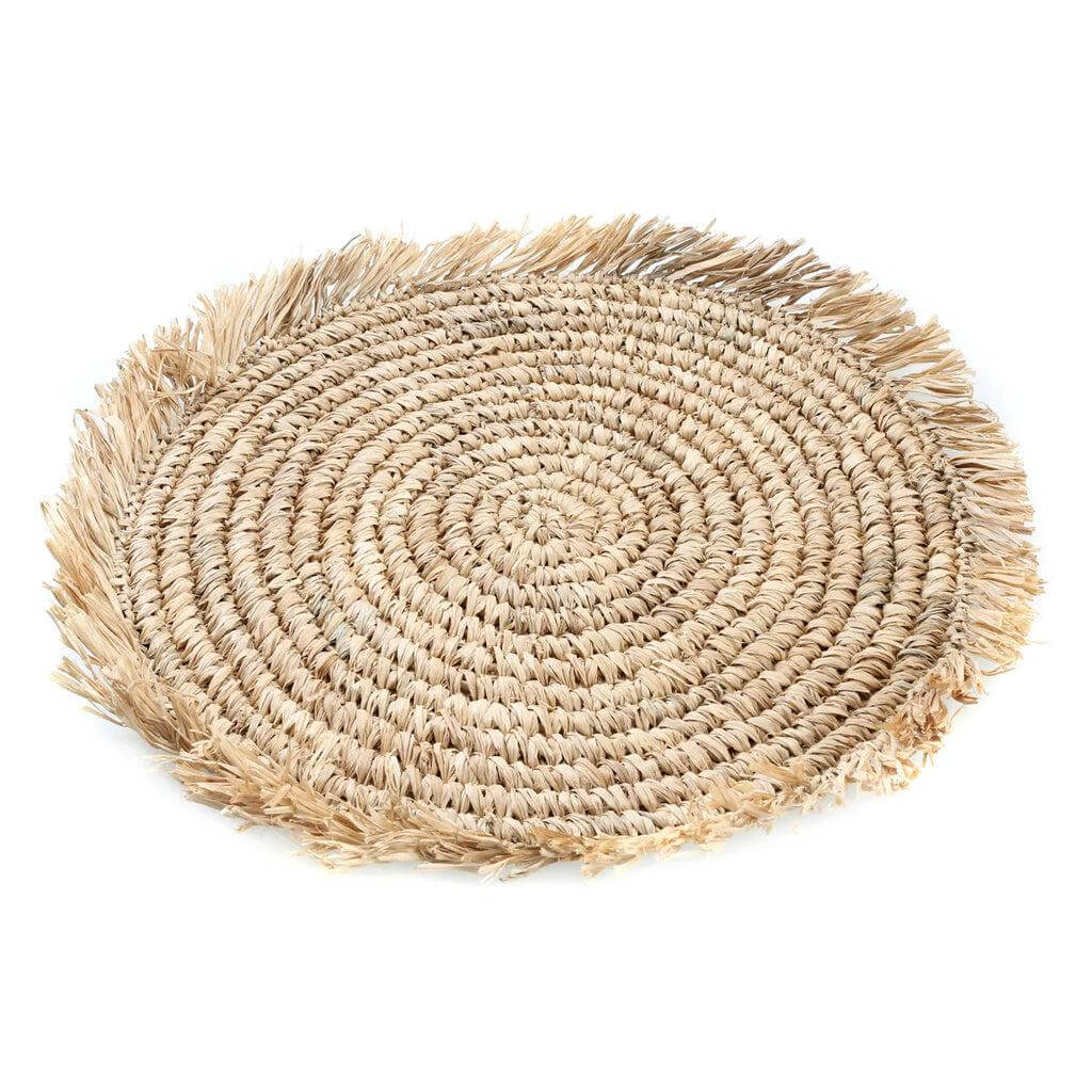 Alt Tag: Handgeweven rotan en raffia placemat met kwastjes, omgeven door kauri schelpen, voegt een tropisch tintje toe aan je tafel.