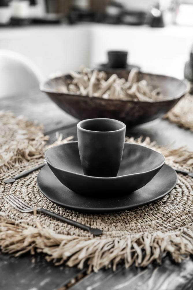 Elegante tafelsetting met handgeweven raffia placemat en zwart serviesgoed voor een tropische sfeer.