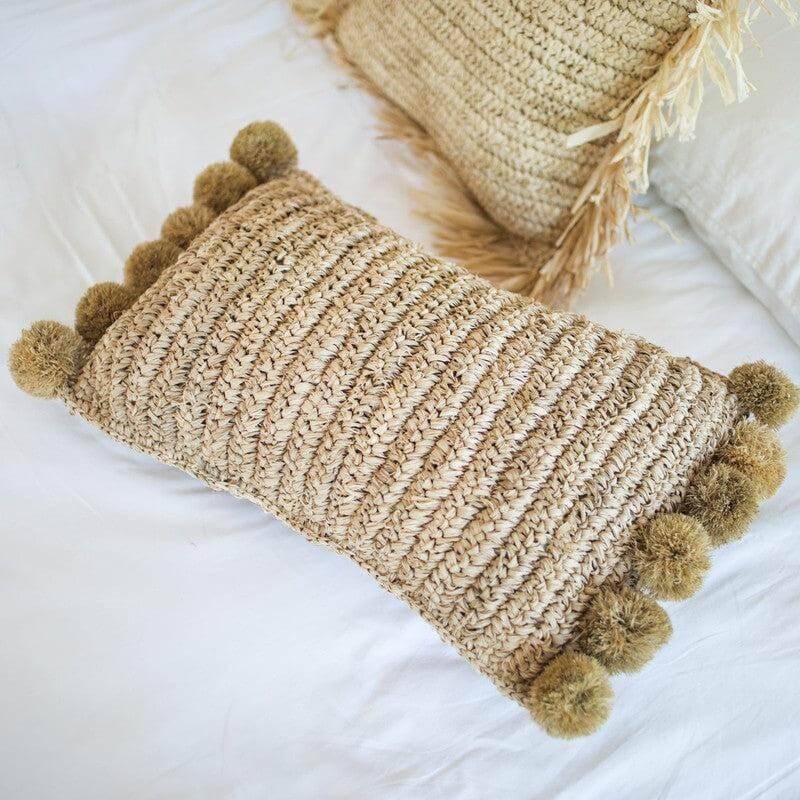 Decoratief rechthoekig sierkussen van raffia met pompons, 50x30 cm, op een wit laken. Handgemaakt, natuurlijk en eerlijk.