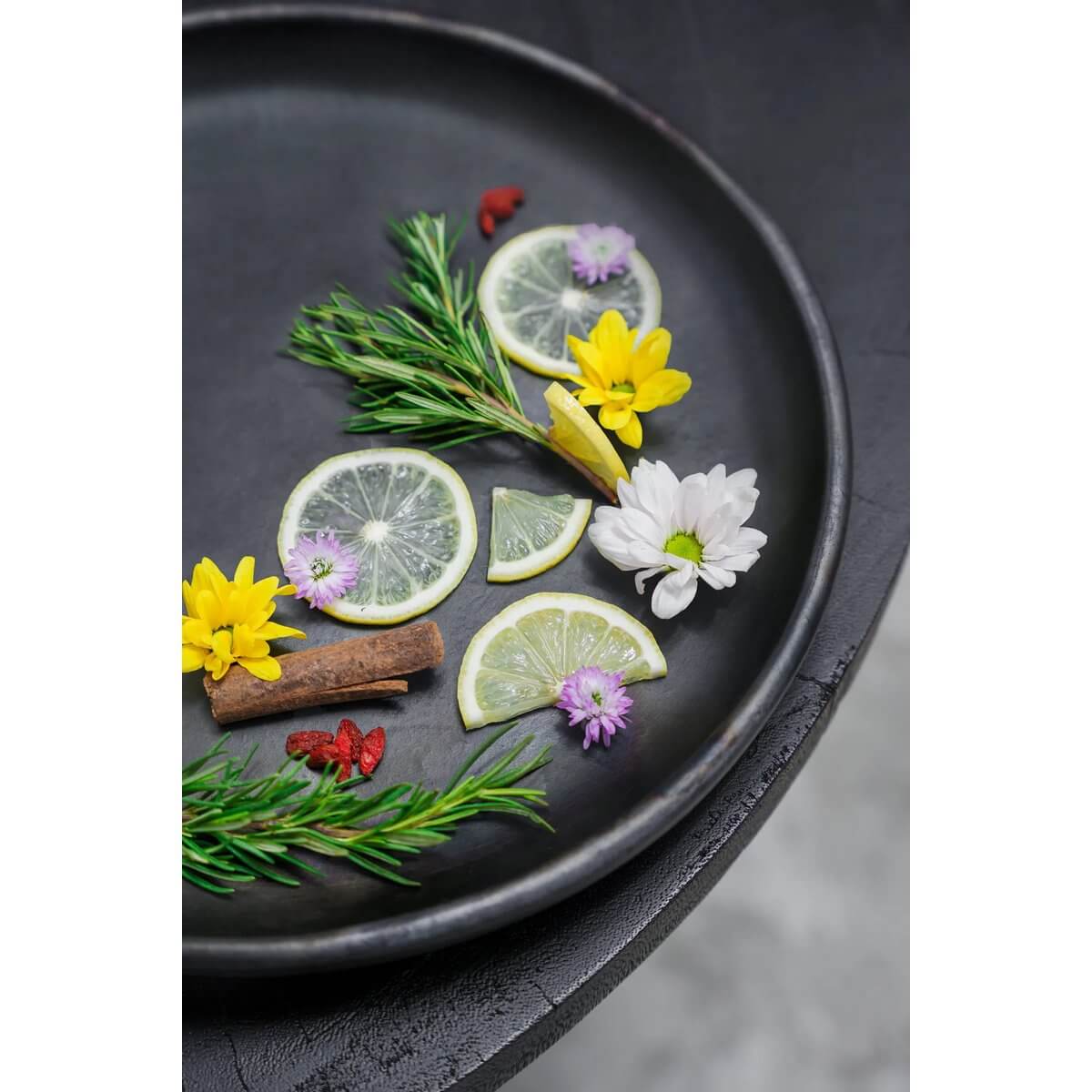 Black ceramic plate with lemon slices, fresh herbs, and flowers, creating a bohemian look. The Burned Classic Plate by Bazar Bizar.