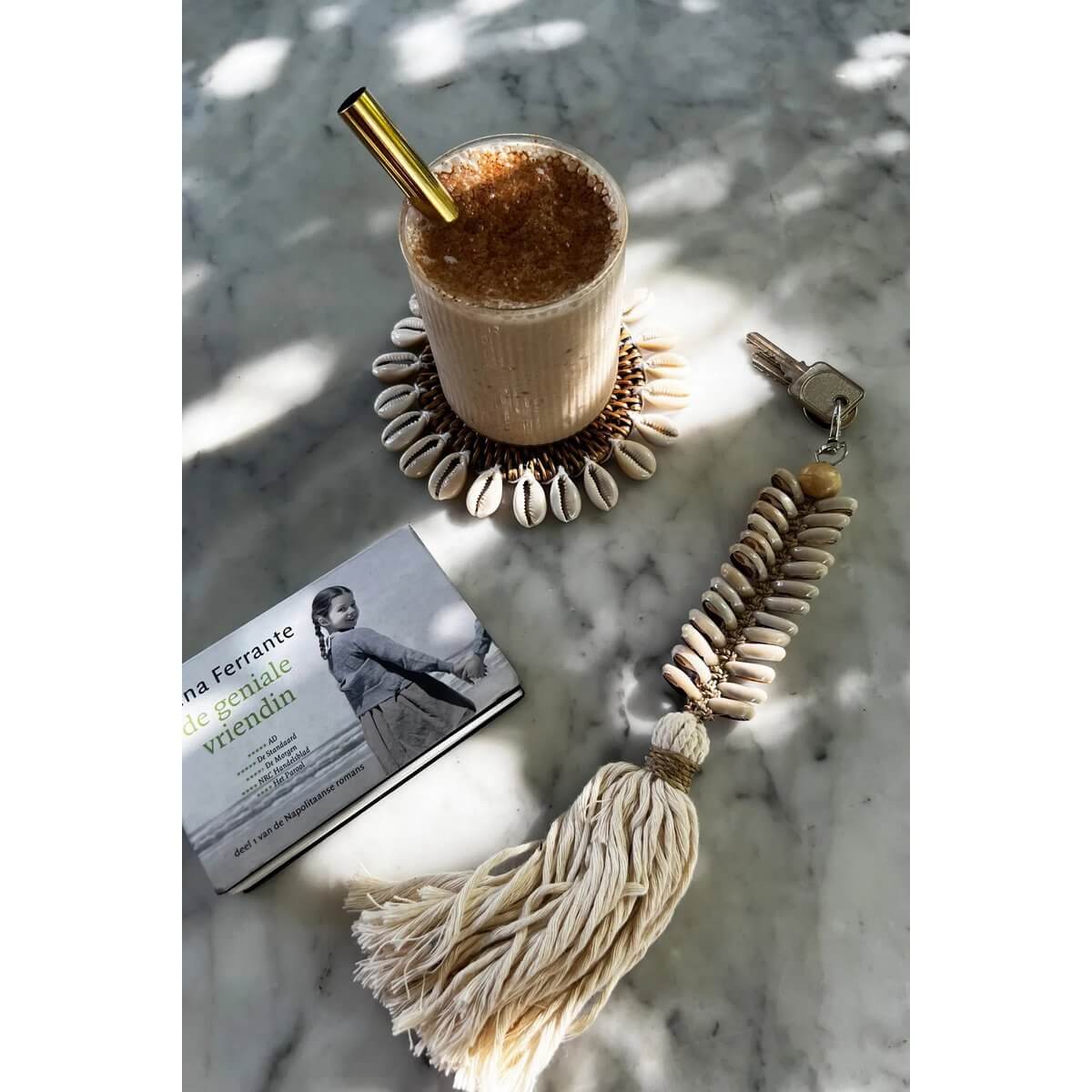 Bohemian vibe: handwoven rattan shell coaster with shells, drink, book, and tassel on a marble surface.