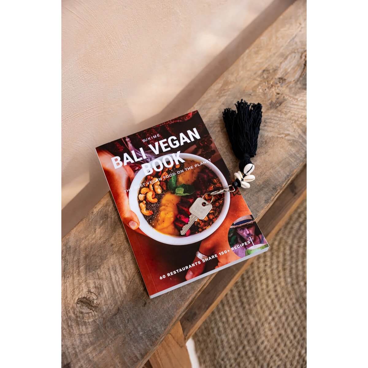 Bali Vegan Book next to Bohemian Cowrie Tassel Keychain with black tassels and shells on a wooden table.