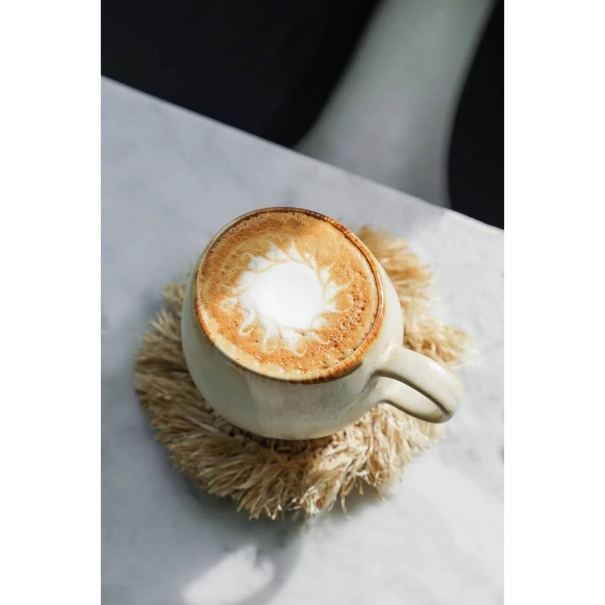 Bohemian Fringe Raffia Coaster in natural tones with a cappuccino mug on a marble table, home decor accent by Bazar Bizar Living.
