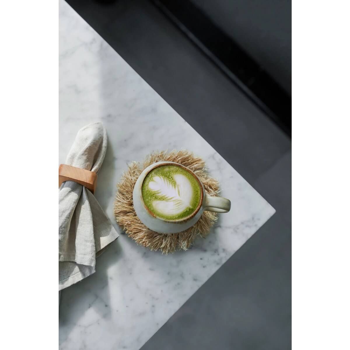 Top view of a bohemian latte on The Fringe Raffia Coaster in natural, beside a cloth napkin, perfect for stylish home decor.