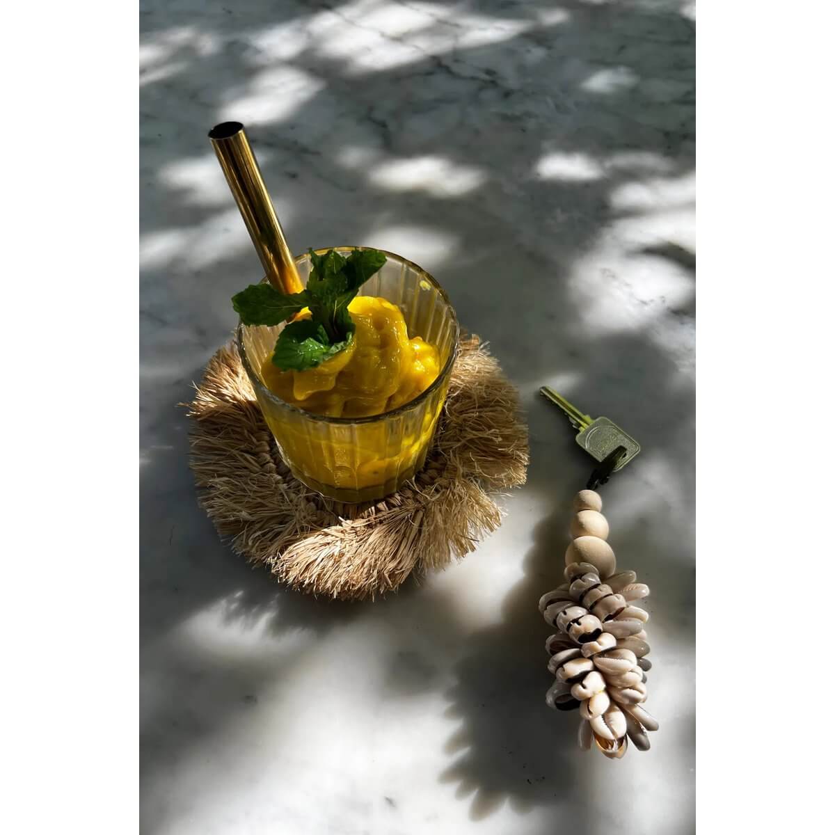 Glass of mango smoothie on a bohemian fringe raffia coaster with metal straw and seashell keychain on marble table.
