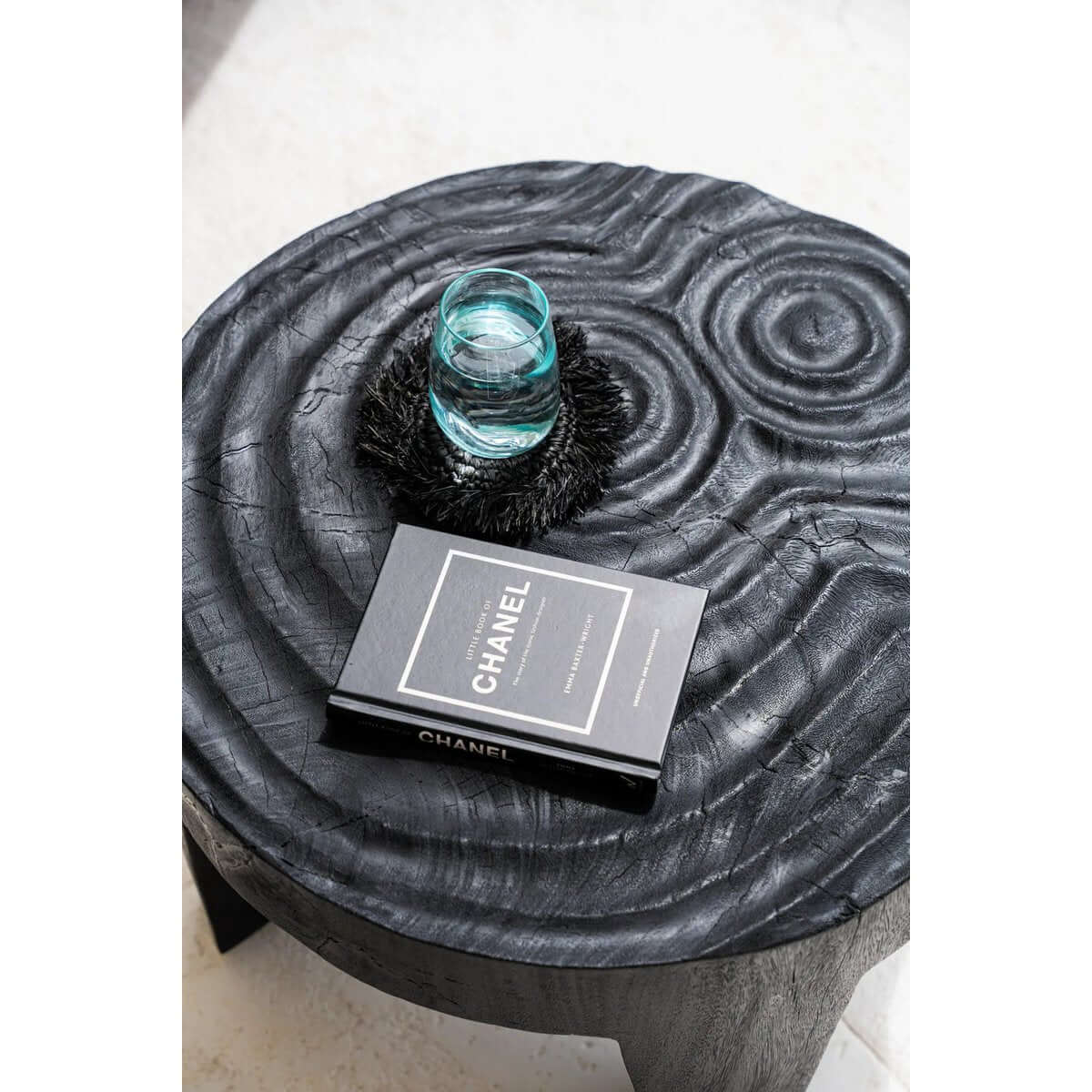 Bohemian-style black raffia coaster on wooden table with a glass cup and Chanel book; perfect for eclectic home decor.