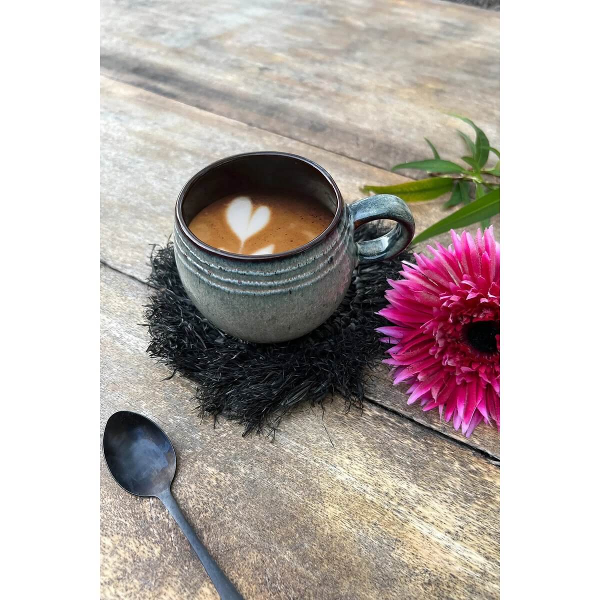Cozy espresso cup on a black raffia coaster with a vibrant pink flower, perfect for a bohemian table setting.