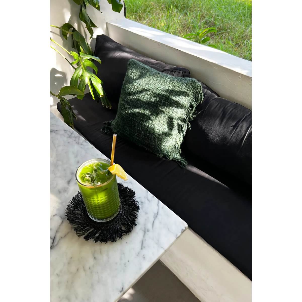 Bohemian style terrace with green smoothie on The Fringe Raffia Coaster - Zwart, featuring a cozy black and green pillow setup.